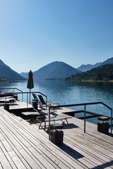 An empty hotel terrace with sun loungers on the shore of a quiet and peaceful alpine lake surrounded by mountains. 