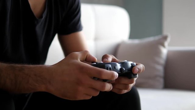 Young man playing video game at home. Nervous guy holding play station in hand. Aggressive gamer sitting on couch with joystick.