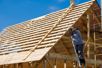 Senior gray-haired builder collects the frame of a wooden country house standing on the stairs against the blue sky. The physical activity of the elderly. Eco-friendly housing made of wood.