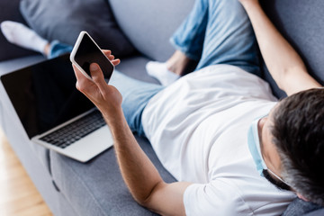 Selective focus of man in medical mask holding smartphone with blank screen near laptop on couch