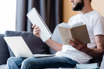 Cropped view of freelancer working with laptop, book and notebook near medical mask in living room