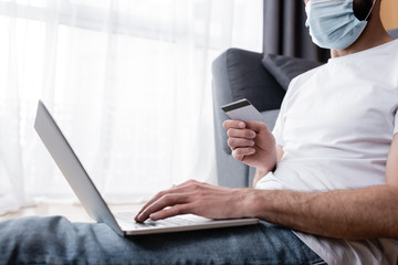 Cropped view of man in medical mask using laptop and credit card at home
