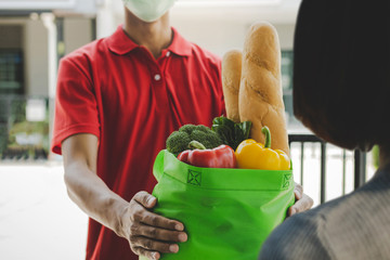 food delivery service man with protection face mask in red uniform holding fresh food set bag to customer at door home, express delivery, quarantine, virus outbreak, takeaway food delivery concept