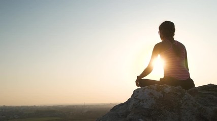 Young relaxed woman sitting outdoors on a big stone enjoying warm summer day. Girl meditating and relaxing on nature at sunset.