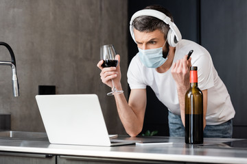 Pensive man in headphones and medical mask holding usb flash drive and glass of wine near laptop in kitchen