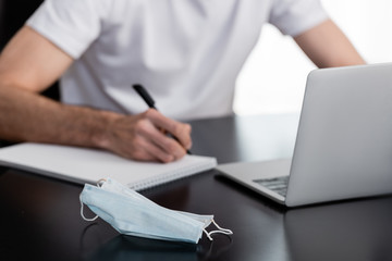 Selective focus of medical mask on table near freelancer working with laptop and notebook at home