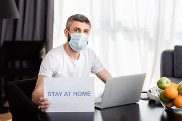 Fototapeta na wymiar Freelancer in medical mask holding card with stay at home lettering near laptop and fruits on table