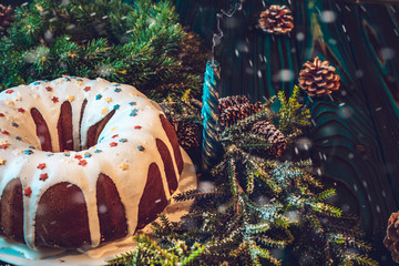 A large festive cupcake decorated with white icing and pastry topping, a blue extinct candle, Christmas tree branches and cones on a blue wooden background. The effect of snowfall.
