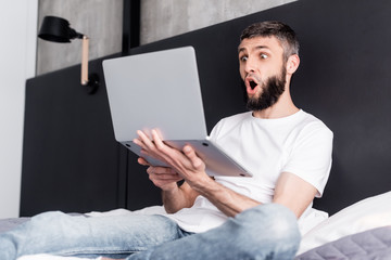 Selective focus of shocked man holding laptop on bed