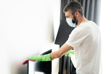 Fototapeta premium Side view of man in medical mask cleaning tv with rag and detergent in living room