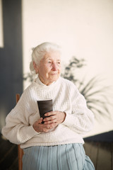 Grandmother is sitting in the chair and drinking tea at home. Beautiful old woman portrait. Grandmother with grey hair is looking into the window and smiling. 