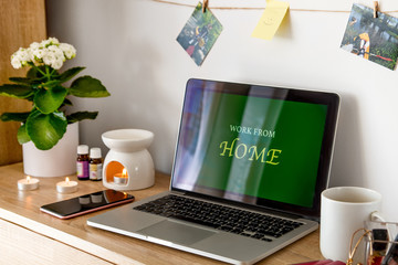 The laptop is on the table. Near the phone, cup, and cosmetics. The screen displays Work from Home. On the wall of the photo.