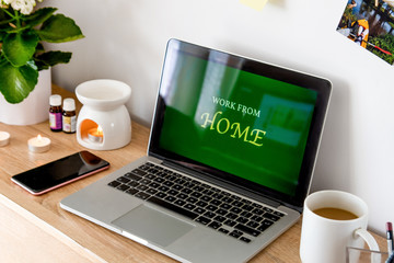 The laptop is on the table. Near the phone, cup, and cosmetics. The screen displays Work from Home. On the wall of the photo.