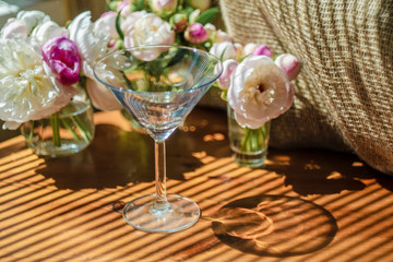 Festive rustic still life with glass empty clear glass surrounded by white flowers on textile background on wooden table. Diagonal striped shadow of the blinds on bright sunny day. Greeting card