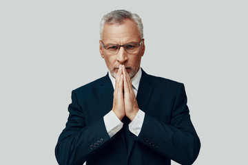 Handsome senior man in full suit looking at camera and keeping hands clasped while standing against grey background