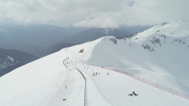 The ski resort. Skiers and snowboarders on the mountain slope. High snow-capped mountains. Krasnaya Polyana, Sochi. Aerial photography video. Snow. Cloudy. Season.