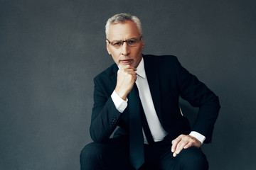 Handsome senior man in full suit looking at camera and keeping hand on chin while sitting against...