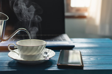 A white cup of coffee with hot smoke in stainless pot, Morning coffee time with notebook computer and work equipment, Normal practice by working at home during covid-19 outbreak around the world.