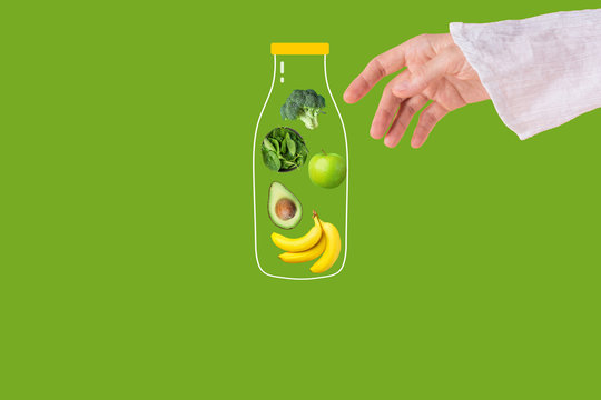 Young Caucasian woman reaching with hand for glass bottle with detox smoothie ingredients fresh raw spinach apples bananas avocado broccoli on green background. Healthy plant based diet