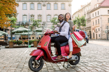 Fototapeta na wymiar Young beautiful multiracial couple, African guy and Caucasian girl, riding on red motorbike on the city street, happy woman holds shopping bags. Summer Europe vacation, traveling concept