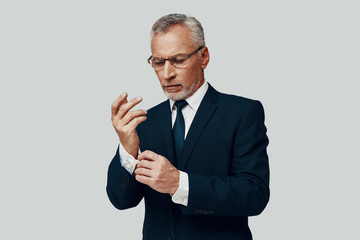 Handsome senior man in full suit adjusting sleeve while standing against grey background
