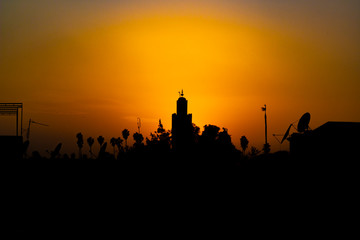 Fototapeta premium Orange sunset above the minaret of the mosque of Kutubiyya, Marrakesh. Black silhouettes by sunset showing minaret, numerous television antennas and palm tree. Muslim evening prayer time