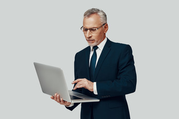 Handsome senior man in full suit using laptop while standing against grey background