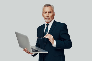 Handsome senior man in full suit using laptop and looking at camera while standing against grey background