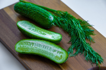 Two halves of cucumber and one whole cucumber are laying on a wooden cutting board together with fresh green dill, closeup macro view