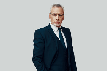 Handsome senior man in full suit looking away while standing against grey background