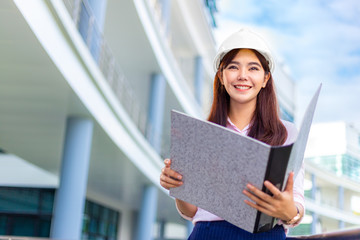 Happy young attractive female Asian engineer wearing hard hat holding her large size blueprint folder while standing outside of office building