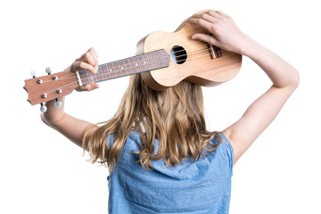 Young, blond girl in blue T-shirt is holding and playing light brown ukulele instrument.