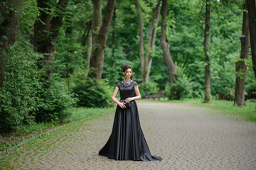 Young beautiful woman posing in a black dress in a park. Holding a handbag in his hands.