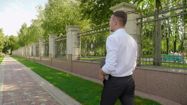 Businessman with laptop in hand walks through the park and looks around. Man freelancer in white shirt and trousers goes to meeting with client. 4K footage