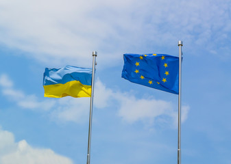 The flag of Ukraine and the flag of the European Union wave in the wind next to each other on flagpoles against the sky.