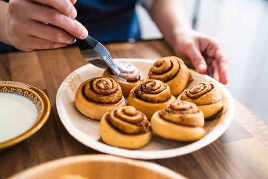 Close up of a Woman's hand painting freshly baked homemade cinnamon rolls with a delicious cream on a wooden counter in her kitchen.