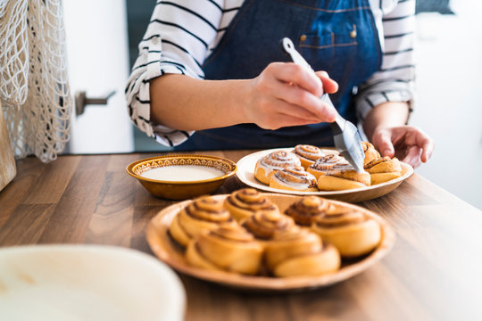 Woman painting freshly baked homemade cinnamon rolls with a delicious cream on a wooden counter in her kitchen.