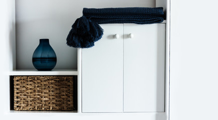 Cozy home interior decor: pillow, plaid, blue vase,  wicker box on a white shelf in the room. The quarantine concept of stay home, distance home education background.