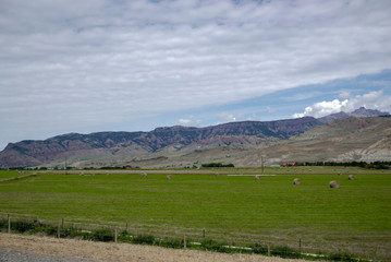 Fototapeta na wymiar Green field with small hay bale there and mountains background cloudy sky with grey blue shade. Road to Yellowstone National Park in Wyoming state, picture for travel blog with copy space.