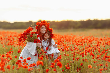Little girl with redhead mother in white dresses and wreathes poses with bouquet of poppies at summer sunset on poppy field