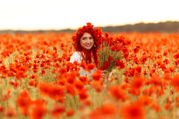 Beautiful redhead smiling woman in wreath with bouquet of poppies on green field