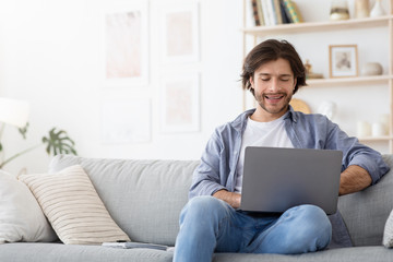 Cheerful guy sitting on sofa at home, using laptop