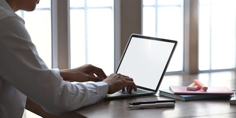 Photo of smart man working as accountant while sitting and working with white blank screen computer laptop at the wooden working desk that surrounded by office equipment over windows as background.