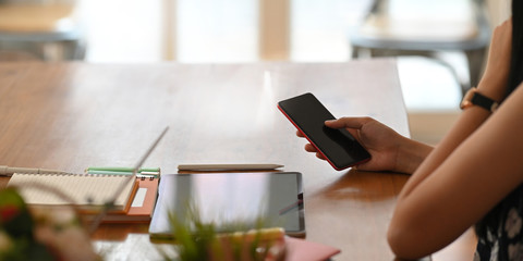 Cropped image of young entrepreneur woman using smartphone while sitting in front her computer tablet that putting on wooden working desk over comfortable sitting room windows as background.