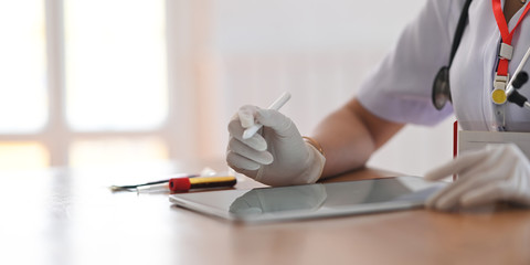 Cropped image of woman working as doctor writing on computer tablet by stylus pen while sitting at the doctor working desk over orderly workplace as background.