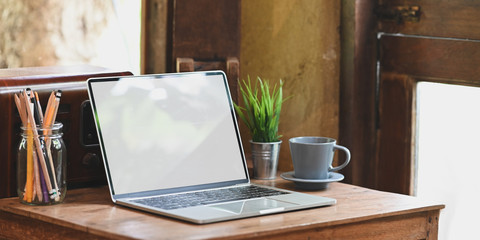 Laptop with white blank screen putting on old wooden desk together with potted plant, coffee cup and pencils in glass vase over comfortable living room as background.