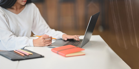 Cropped image of creative woman in white cotton shirt working with computer laptop while holding a pencil and sitting at white working desk that surrounded by notebook, pencils and computer tablet.