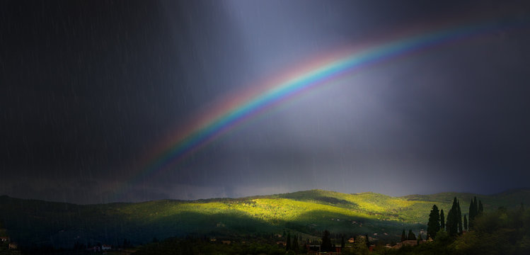 Rain, sunbeams and Rainbow in dramatic dark sky over green hillside;  beautiful summer countryside landscape