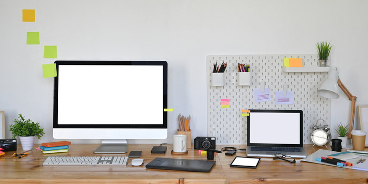 Photo of computer laptop and computer monitor with white blank screen putting on wooden working desk that surrounded by graphic designer equipment over white living room wall as background.