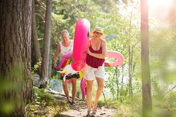 Elderly couple walking a forest path along the seashore holding giant inflatable flamingo and unicorn. Funny active pensioners enjoying summer vacation on the beach in Northern Europe
